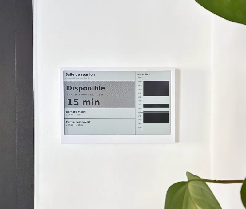 Écran e-ink monté au mur devant une salle de réunion, affichant sa disponibilité et l’agenda du jour